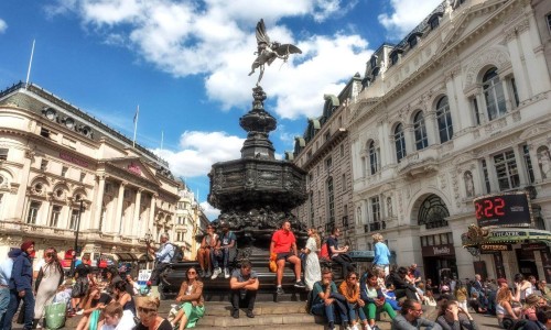 Tourists sitting on the steps of the Shaftesbury Memorial Fountain and statue of Greek god Anteros, Piccadilly Circus, London’s West End.